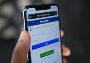 Picture of a hand holding an iPhone with the Facebook app pulled up. The Facebook login screen is showing.