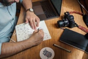 Person drawing cursive letter "A" on wooden desk, next to a camera, pen, and notebook
